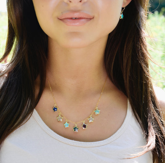 A woman wearing a handmade 14k gold filled necklace of seven stars. Each star is a different gemstone. Stones include: Lapis lazuli, rainbow moonstone, turquoise, labradorite, amethyst, rose quartz, and amazonite.