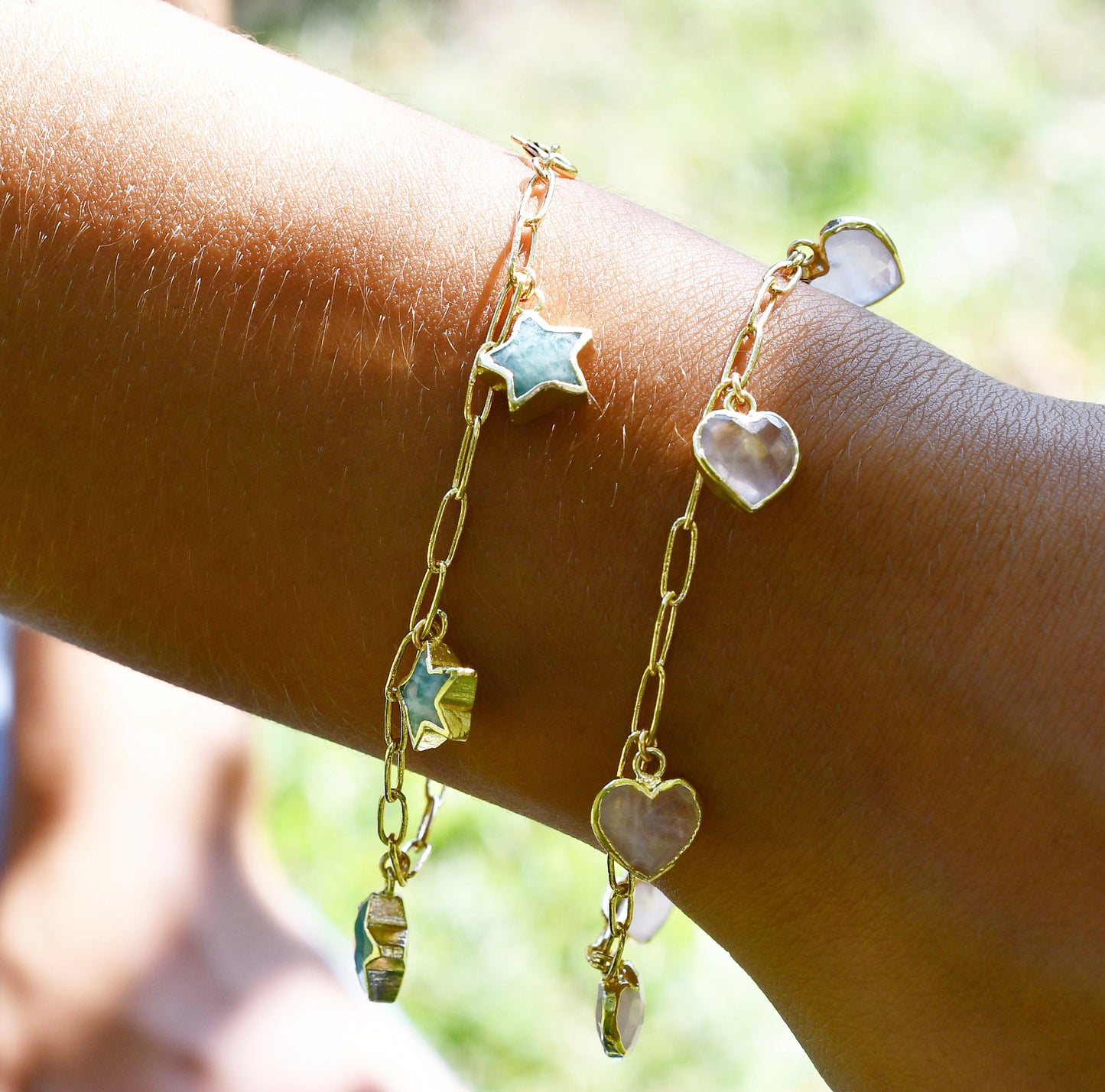 An amazonite star bracelet layered with a pink rose quartz heart bracelet. Both are on gold dainty paperclip chains. Modeled image.