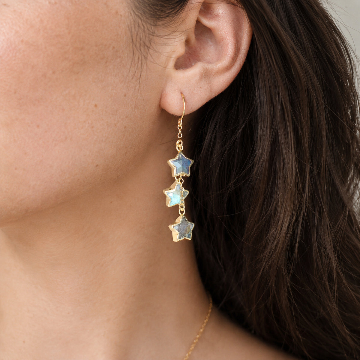 A woman wearing a pair of dangle celestial earrings made with dainty blue and green flash labradorite gemstones. Handmade in 14k gold filled or sterling silver.
