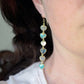 Modeled image. These handmade earrings feature blue and white natural stone hearts bezeled in gold. Each gemstone is a different color and includes: labradorite, larimar, mother of pearl, aquamarine, and rainbow moonstone. Long and light weight they make the perfect romantic gift. They are 3 inches long.