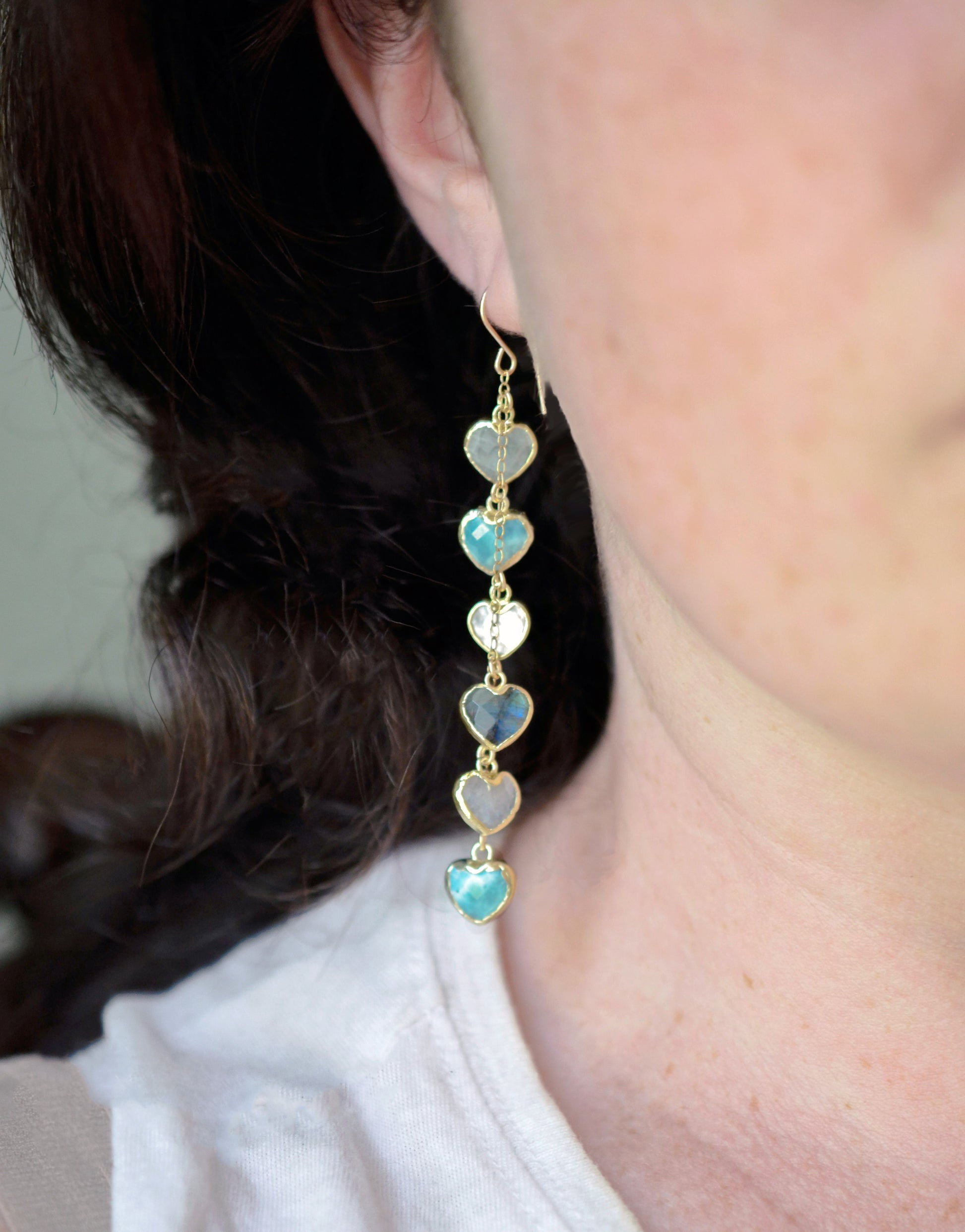 Modeled image. These handmade earrings feature blue and white natural stone hearts bezeled in gold. Each gemstone is a different color and includes: labradorite, larimar, mother of pearl, aquamarine, and rainbow moonstone. Long and light weight they make the perfect romantic gift. They are 3 inches long.