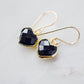 Black onyx heart earrings in gold. The stones are faceted and solid black with gold bezels surrounding their sides. The ear wires are basic hooks.