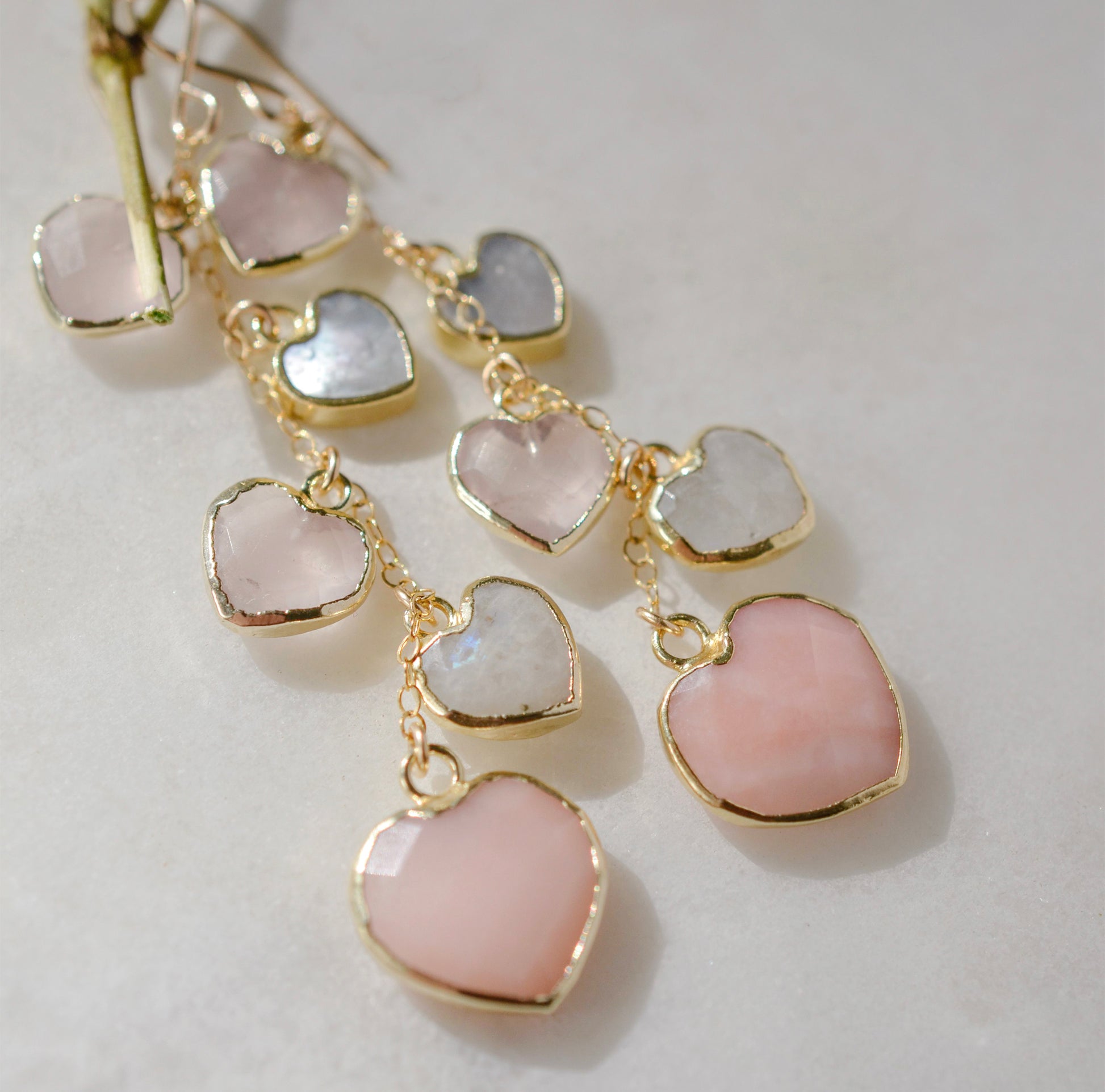 Long pink stone heart earrings in 14k gold filled. There are five heart crystal dangles hanging from a simple chain forming a cluster. The stones include: rose quartz, mother of pearl, pink opal, and rainbow moonstone. They are approximately three inches long.