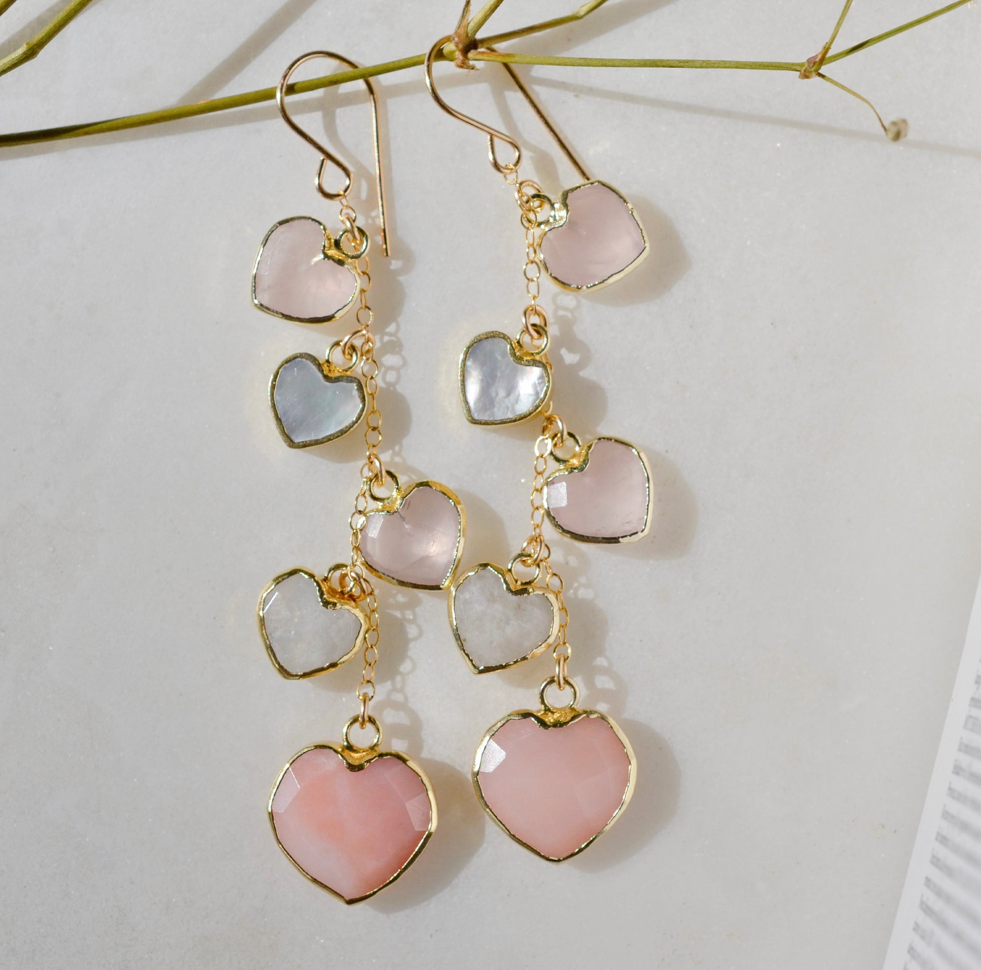 Long pink stone heart earrings in 14k gold filled. There are five heart crystal dangles hanging from a simple chain forming a cluster. The stones include: rose quartz, mother of pearl, pink opal, and rainbow moonstone. They are approximately three inches long.