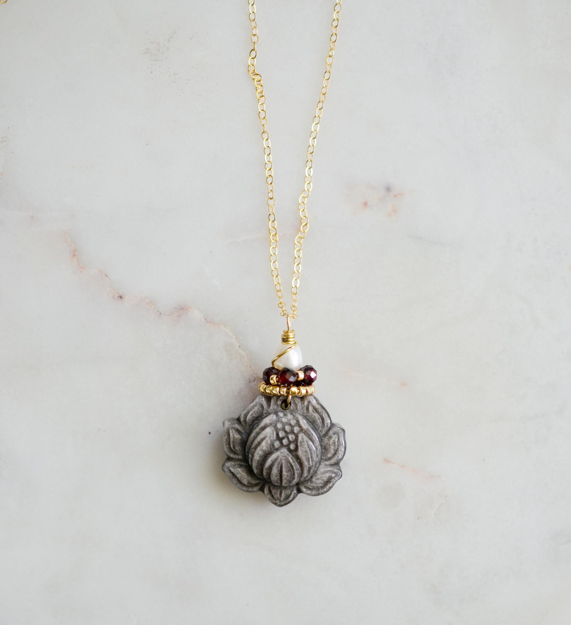 A handmade gemstone pendant featuring a carved obsidian lotus. Garnet, pearl, and gold beads decorate its top. The chain is 14k gold filled.