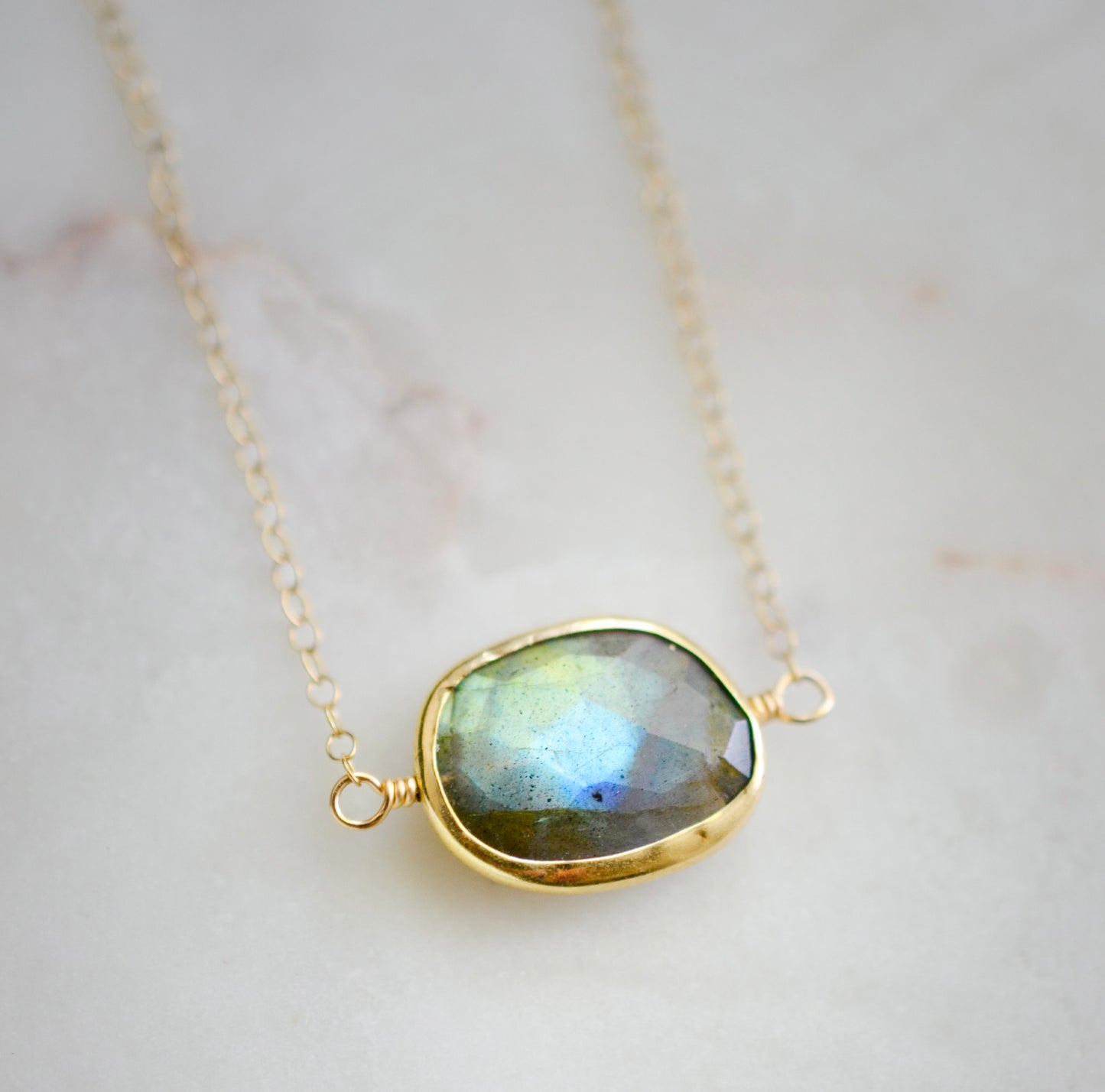 Necklace with a gold-framed Labradorite stone on a light background. The handmade pendant is bezeled in gold and set on a 14k gold filled chain. This necklace is also available in sterling silver. The stone is oval in shape and flashes blue, green, or yellow.
