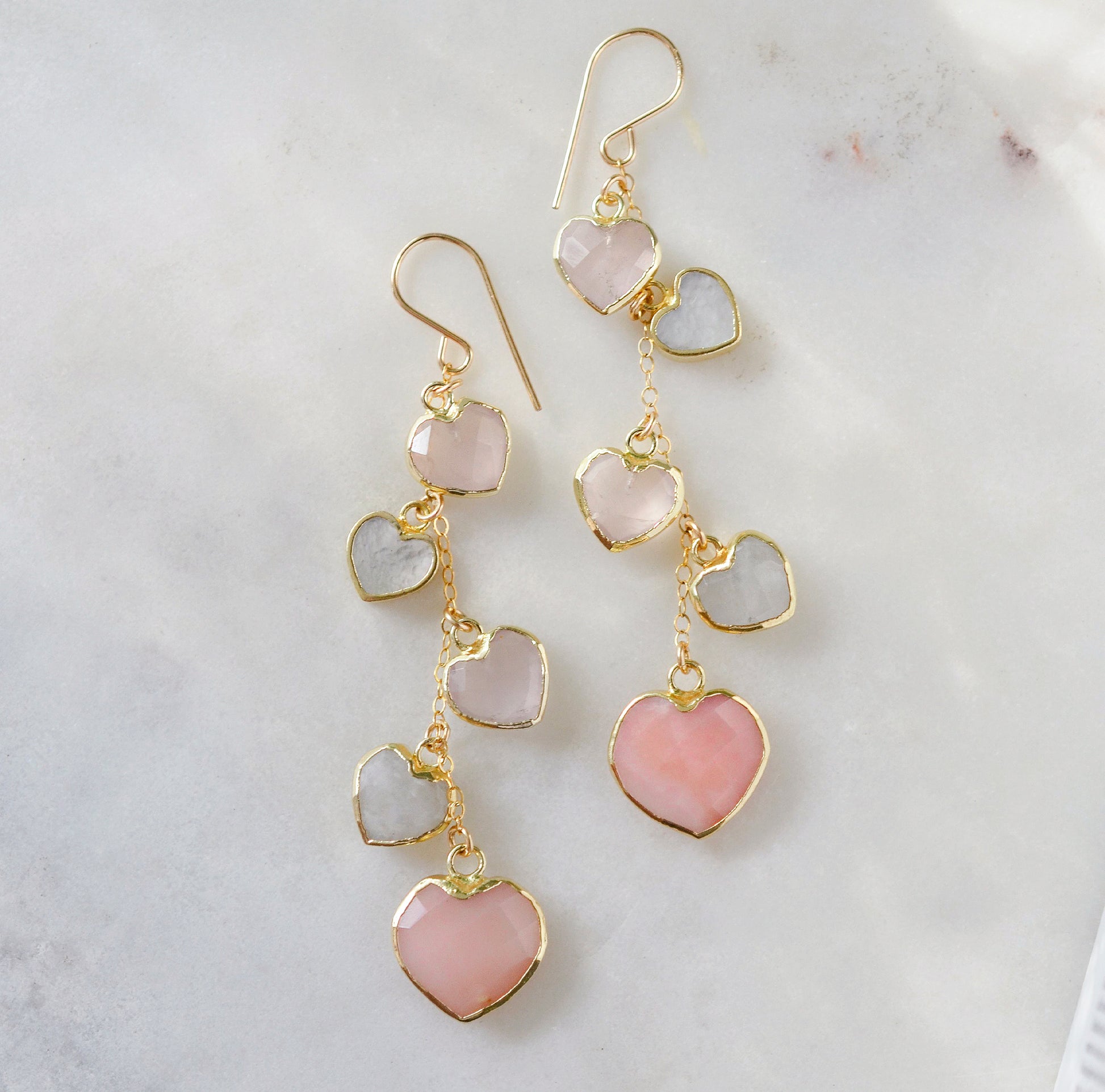 Long pink stone heart earrings in 14k gold filled. There are five heart crystal dangles hanging from a simple chain forming a cluster. The stones include: rose quartz, mother of pearl, pink opal, and rainbow moonstone. They are approximately three inches long.