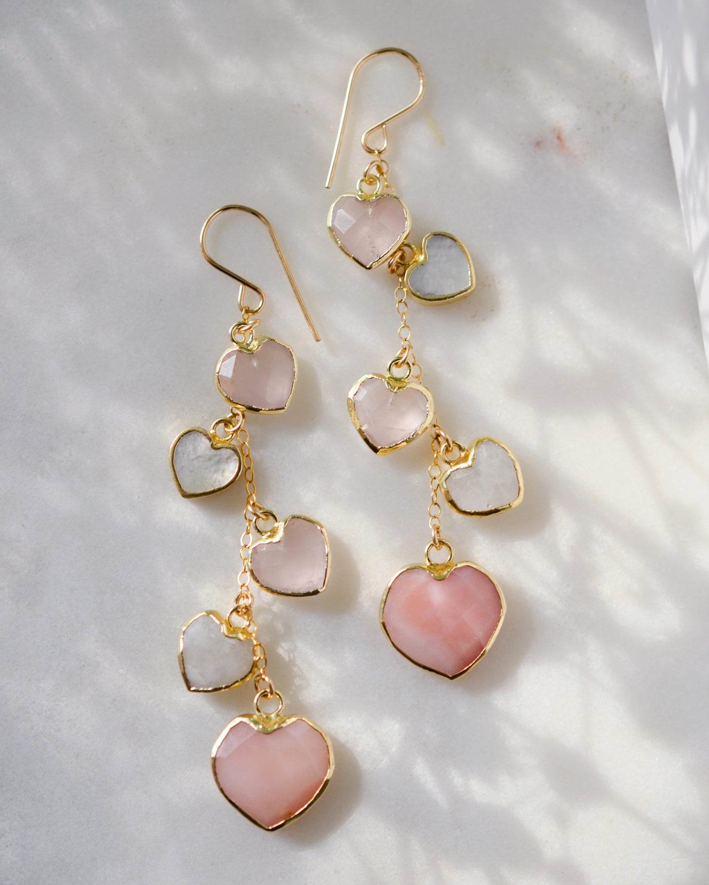 Long pink stone heart earrings in 14k gold filled. There are five heart crystal dangles hanging from a simple chain forming a cluster. The stones include: rose quartz, mother of pearl, pink opal, and rainbow moonstone. They are approximately three inches long.