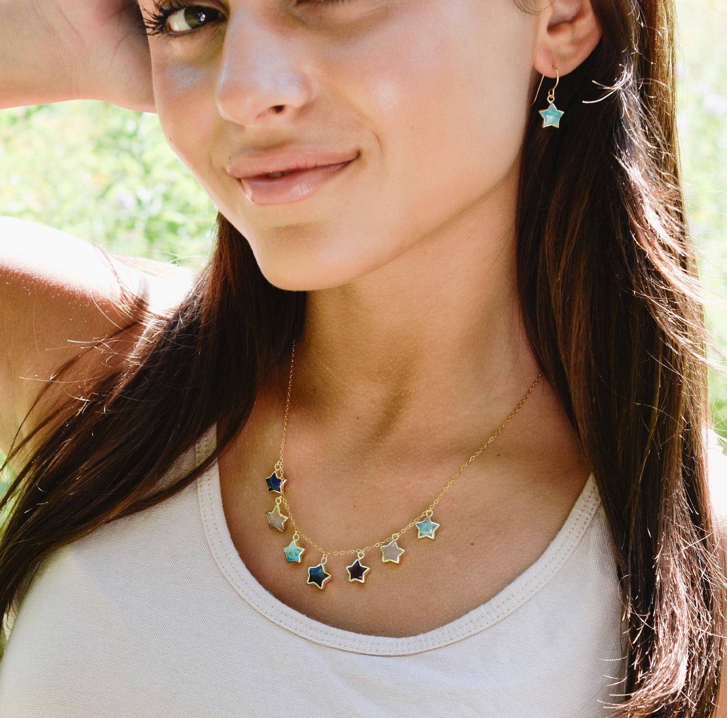 Woman wearing a necklace with star-shaped pendants on a 14k gold filled chain. Each star is a different gemstone. Stones include: Lapis lazuli, rainbow moonstone, turquoise, labradorite, amethyst, rose quartz, and amazonite. This necklace is handmade in Connecticut by Gemnia.