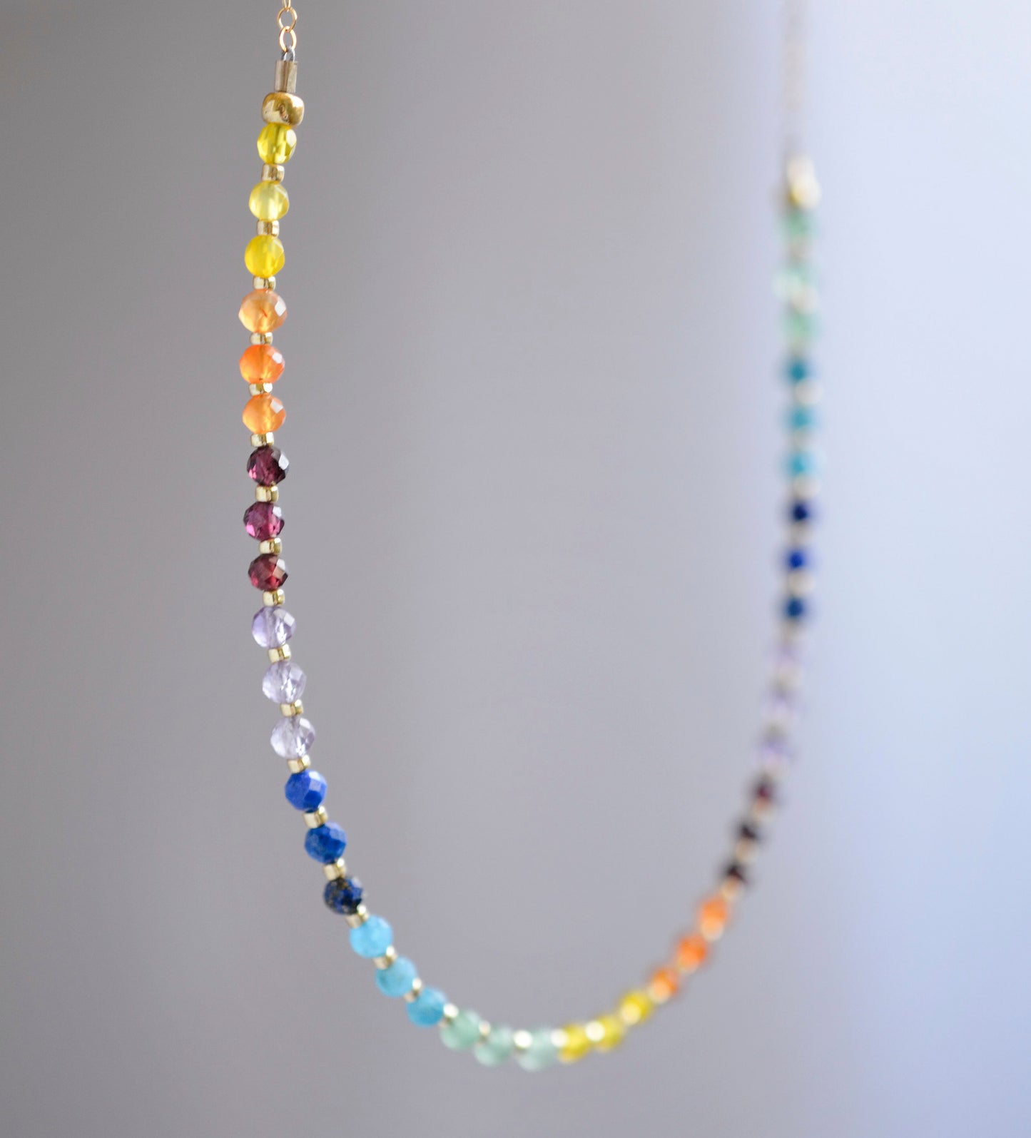 Rainbow chakra gemstone necklace. Crystals are beaded together in this pattern: yellow, orange, red, purple, blue, aqua, green. The gold style is shown. Close up.