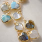 Close up image. These handmade earrings feature blue and white natural stone hearts bezeled in gold. Each gemstone is a different color and includes: labradorite, larimar, mother of pearl, aquamarine, and rainbow moonstone. Long and light weight they make the perfect romantic gift. They are 3 inches long.